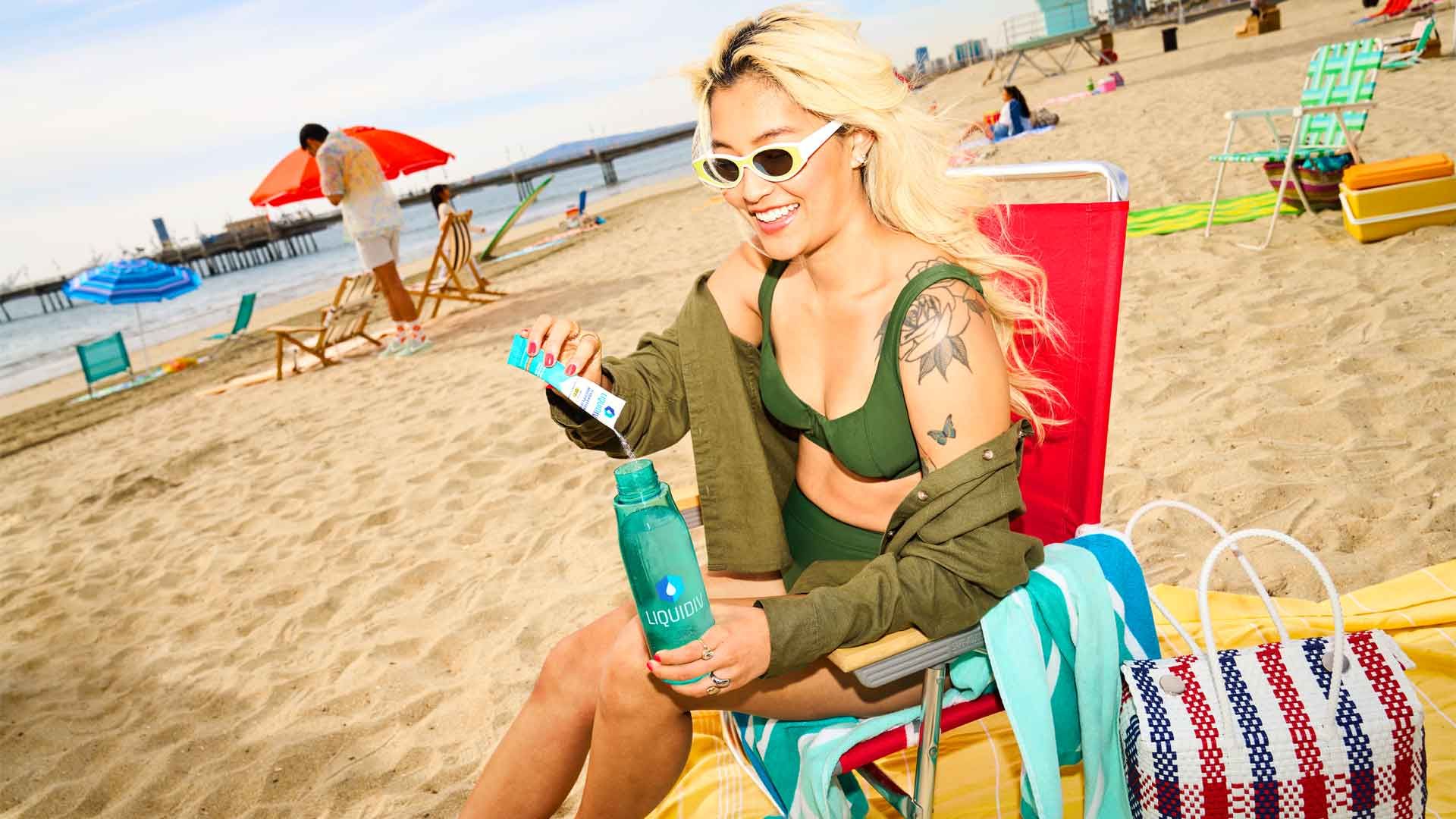 A woman on a beach pours Liquid I.V. powder into a bottle of water to prepare a drink